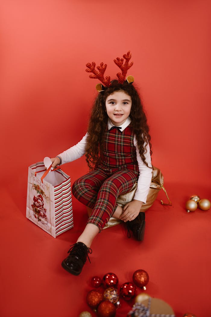 Smiling child wearing reindeer antlers and plaid outfit with a gift bag on a red background.