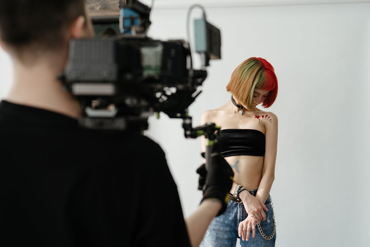 A tattooed woman with colored hair poses for a photoshoot in a tattoo studio while being filmed.